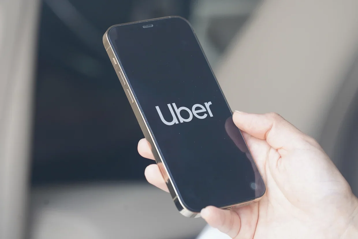 A close-up of a person holding a smartphone displaying the Uber app logo. The phone is held in one hand inside a vehicle.