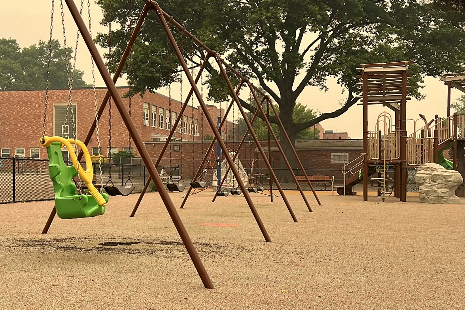 A playground featuring a row of swing sets, with one adaptive swing in the foreground designed for children with disabilities. The swing is green with a yellow harness, contrasting with the standard black swings in the background. The scene includes a climbing structure, benches, and a large tree providing shade. A brick school building is visible in the background, and the area is surrounded by a chain-link fence. The muted lighting suggests an overcast or hazy day.