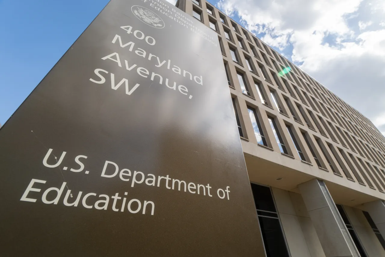 A low-angle view of the U.S. Department of Education building at 400 Maryland Avenue, SW, Washington, D.C., with a brown sign in the foreground displaying the department's name. The sky is partly cloudy with a lens flare visible.