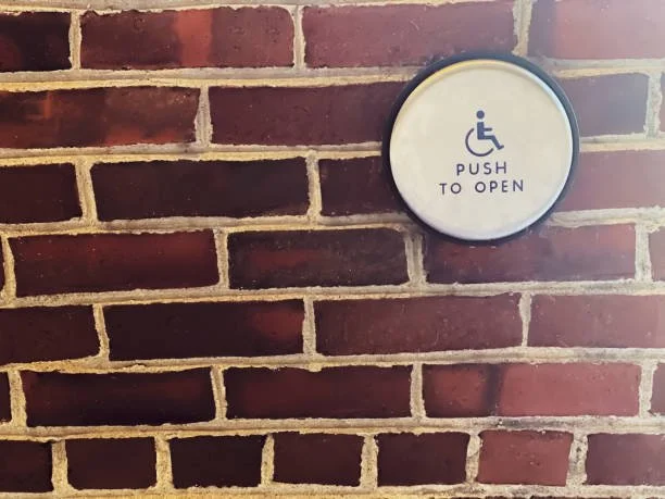 A brick wall with a round button labeled "PUSH TO OPEN" featuring a wheelchair accessibility symbol.