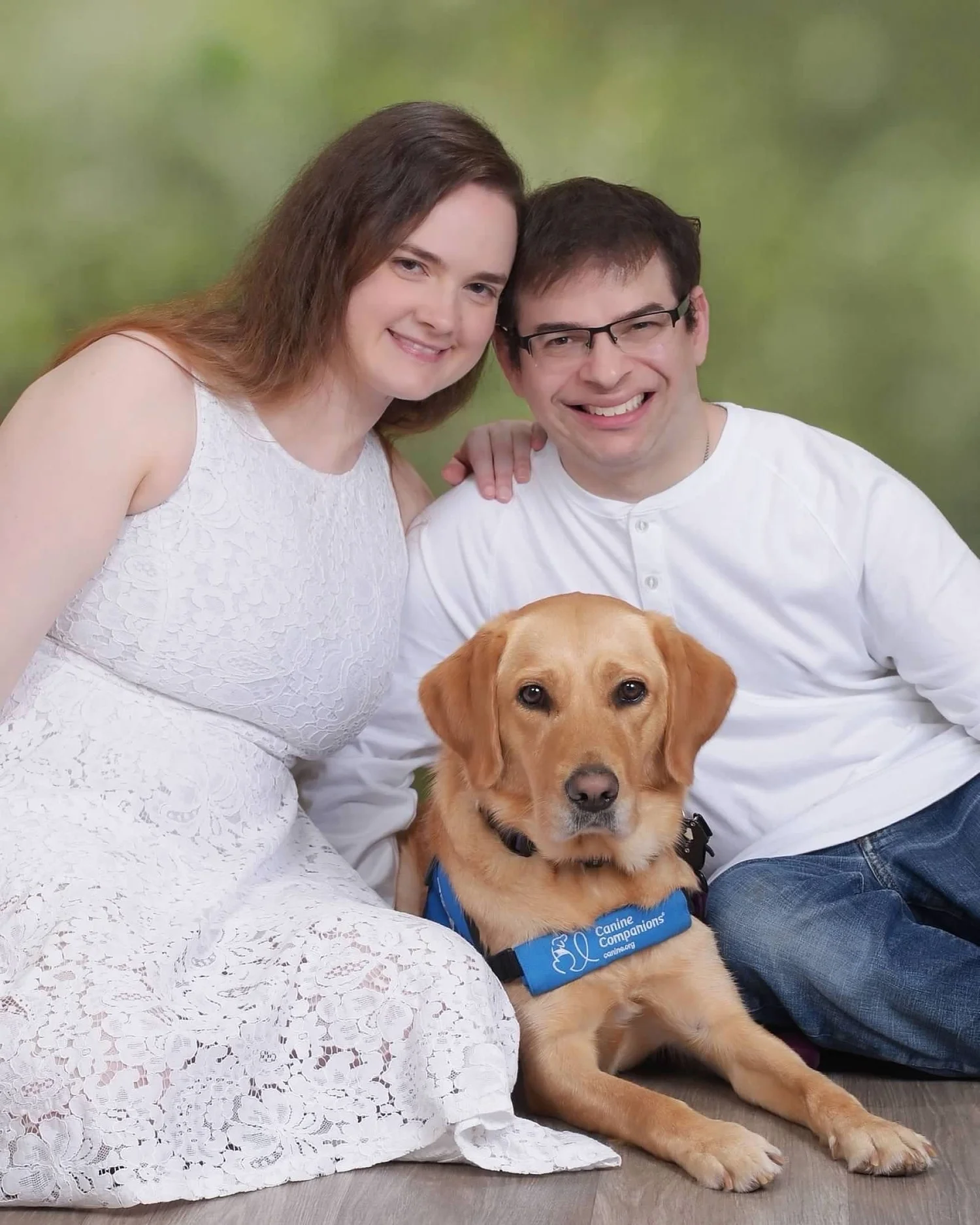 A professional portrait of me, my girlfriend, and my service dog, Lovey. We’re seated on a light wood floor with a soft green backdrop behind us. I’m in a white shirt and jeans; she’s in a white lace dress. Lovey, in her blue Canine Companions vest, lies calmly between us. The photo captures not just a moment, but our bond—grounded in love, advocacy, and pride during Disability Pride Month.
