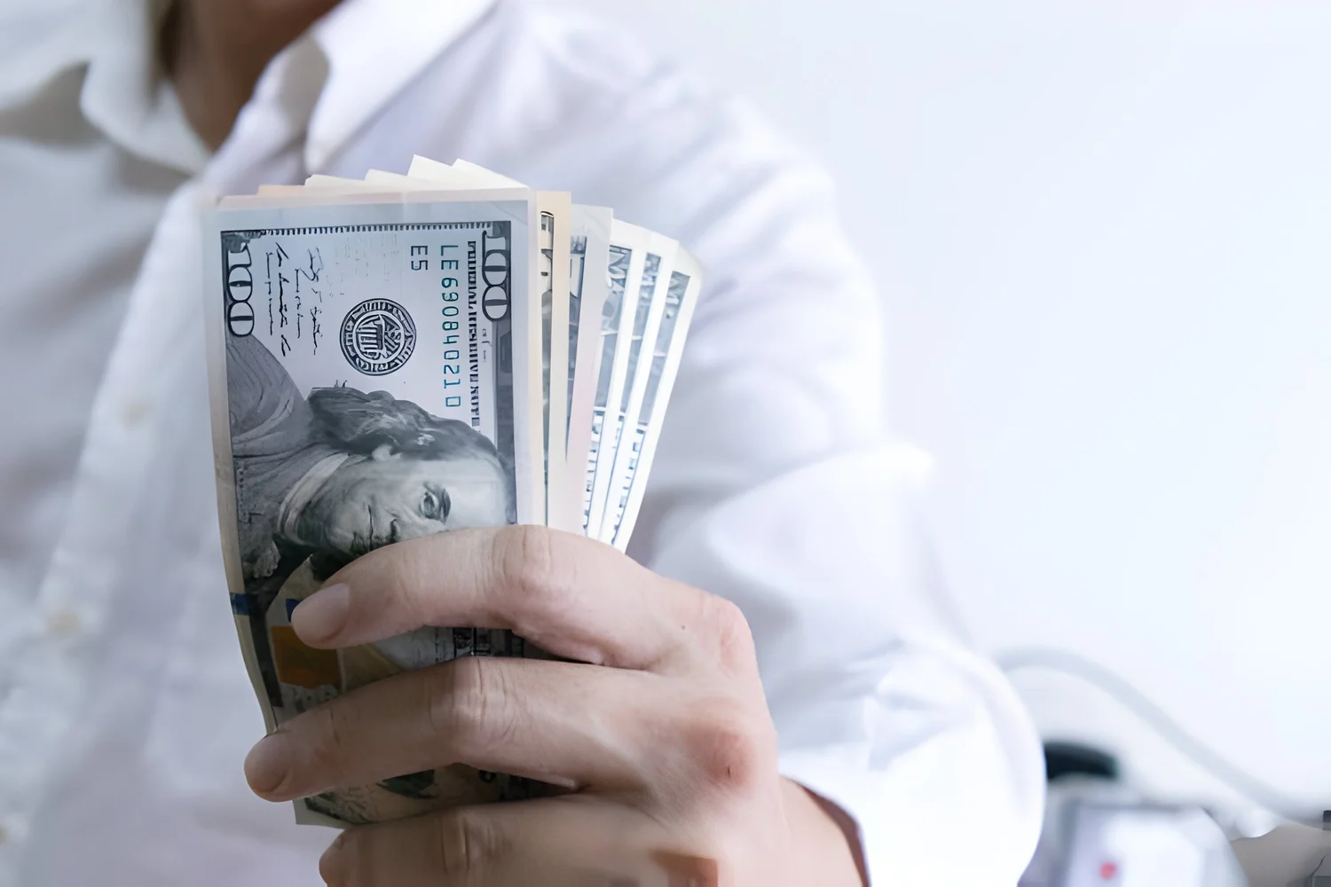 A close-up image of a person in a white shirt holding a fan of hundred-dollar bills in their hand. The focus is on the money, showing several crisp banknotes prominently featuring Benjamin Franklin. The background is blurred, drawing attention to the hand and the cash.