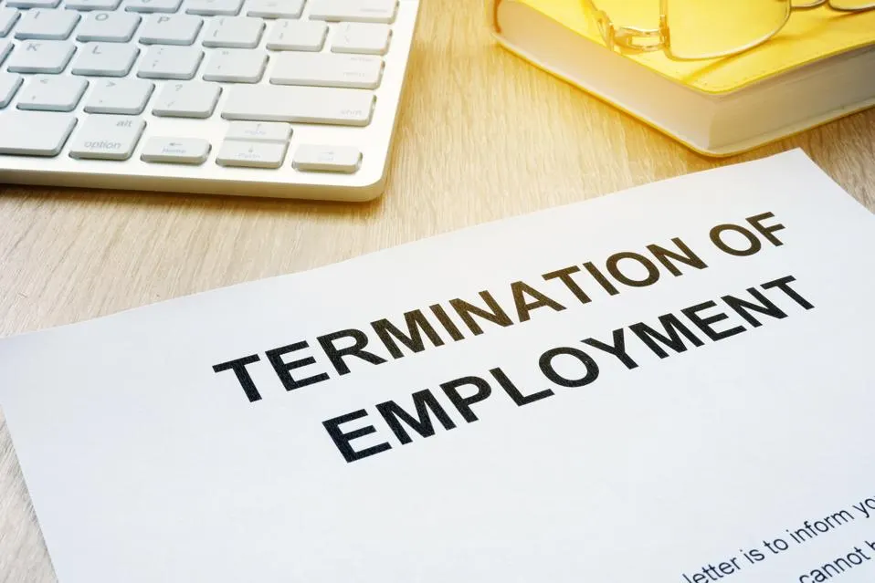 The image shows a close-up of a document titled "Termination of Employment" placed on a wooden desk. To the side, there is a partial view of a keyboard and a yellow-tinted pair of eyeglasses resting on a closed book.