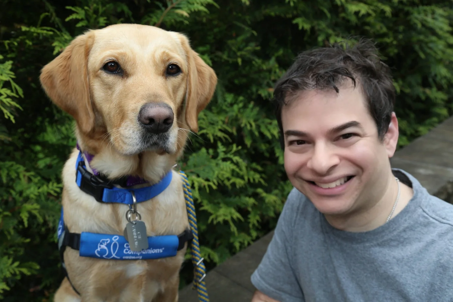 Ryan Honick smiles beside his service dog Lovey, a yellow Lab in a blue Canine Companions vest, with green foliage in the background.