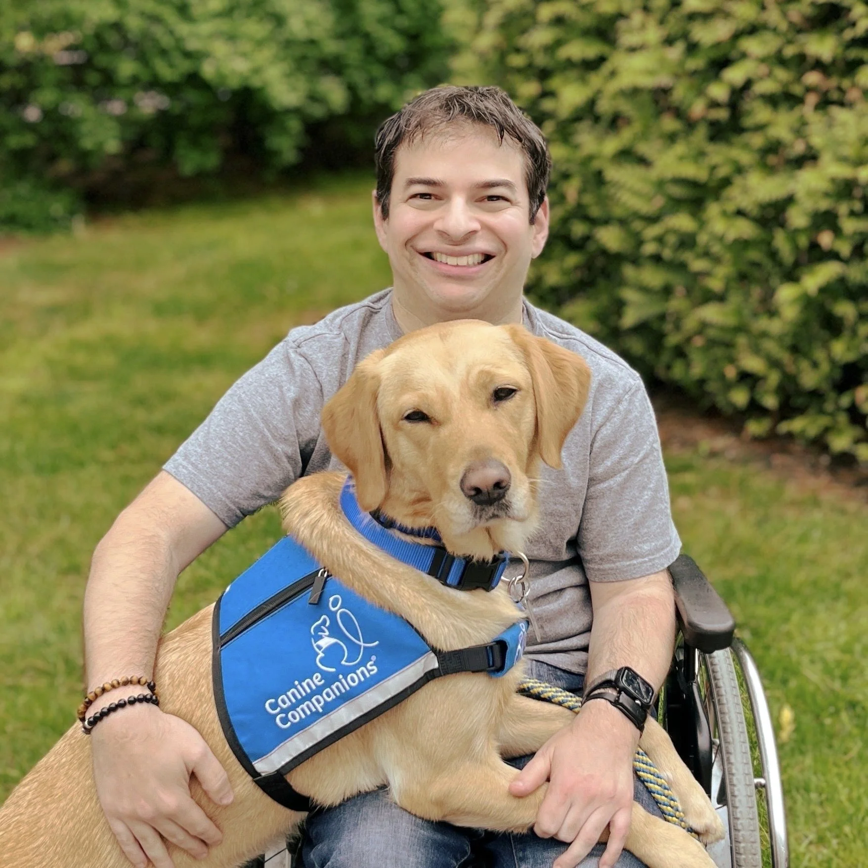 Ryan Honick smiling outdoors with Lovey resting across his lap in her blue service-dog vest